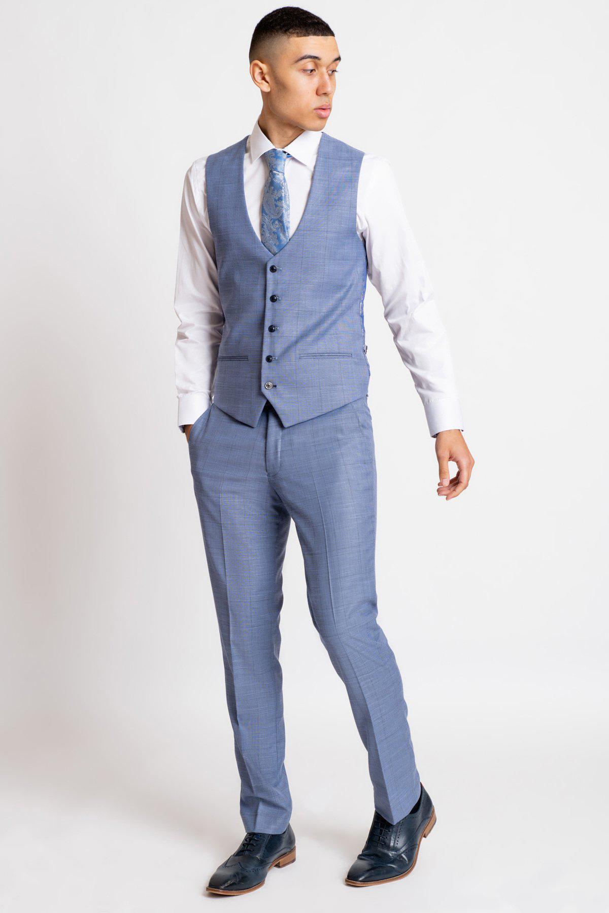 A young man in a Marc Darcy SID Blue Grey Check Single Breasted Waistcoat, white shirt, blue patterned tie, and dark dress shoes stands with hands in pockets, looking sideways against a plain white background.