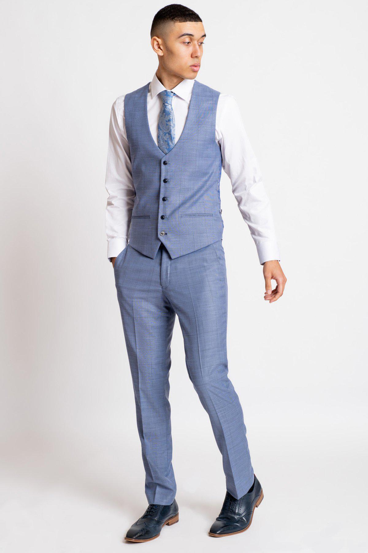 A man stands against a white background wearing marcdarcys SID - Blue Grey Check Trousers, a matching vest, white dress shirt, blue patterned tie, and dark shoes. His hands are in his pockets as he looks to the side.