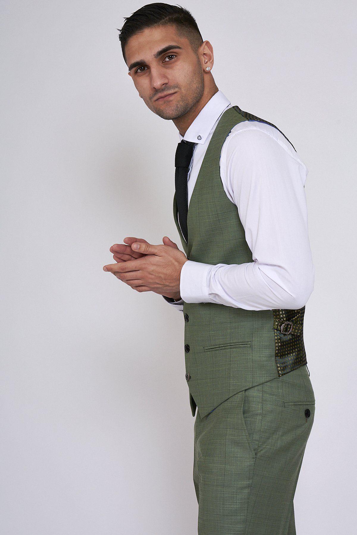 A man wears the Marc Darcy SID Moss Green Check Single Breasted Waistcoat, styled with a white shirt and black tie, standing sideways with hands clasped against a plain light background, looking at the camera.