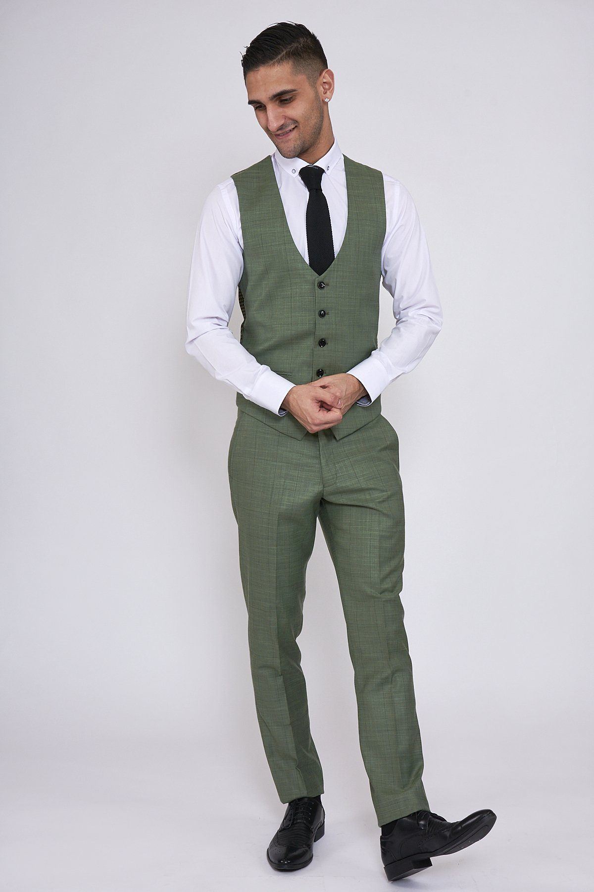A man stands against a plain white background wearing the Marc Darcy SID - Moss Green Check Single Breasted Waistcoat, paired with a matching slim fit suit, white shirt, black tie, and black dress shoes. He smiles down with his hands loosely clasped.