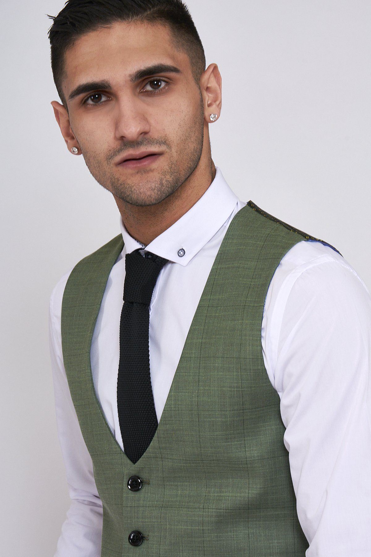 A man with short dark hair and earrings wears the Marc Darcy SID - Moss Green Check Single Breasted Waistcoat over a slim fit white shirt and black tie, standing against a plain background and looking slightly to the side.