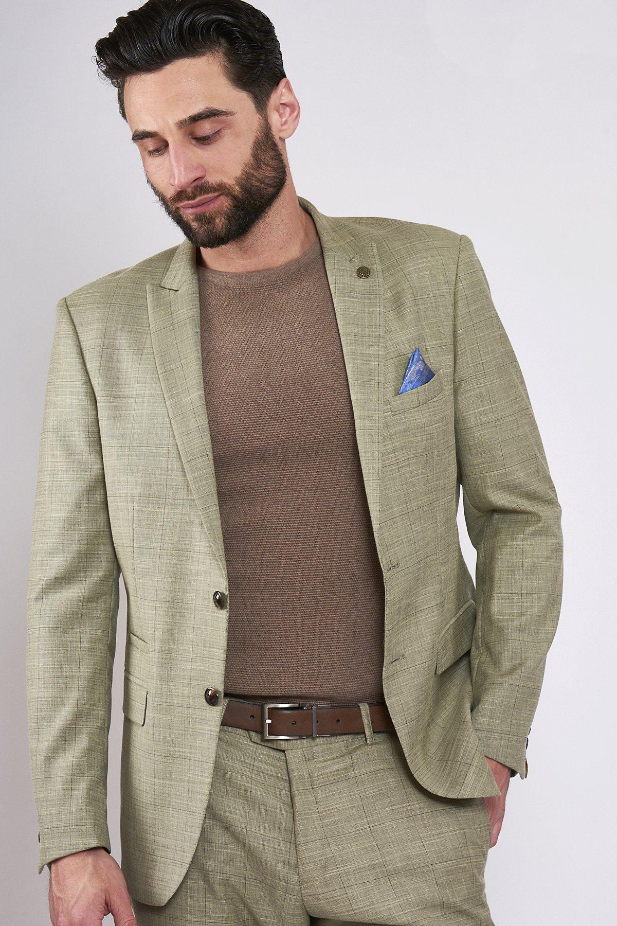 A man with dark hair and beard wears the marcdarcy SID - Mustard Beige Check Blazer, styled with a brown shirt, brown belt, and blue pocket square. He stands against a plain light background, looking downward with a neutral expression.