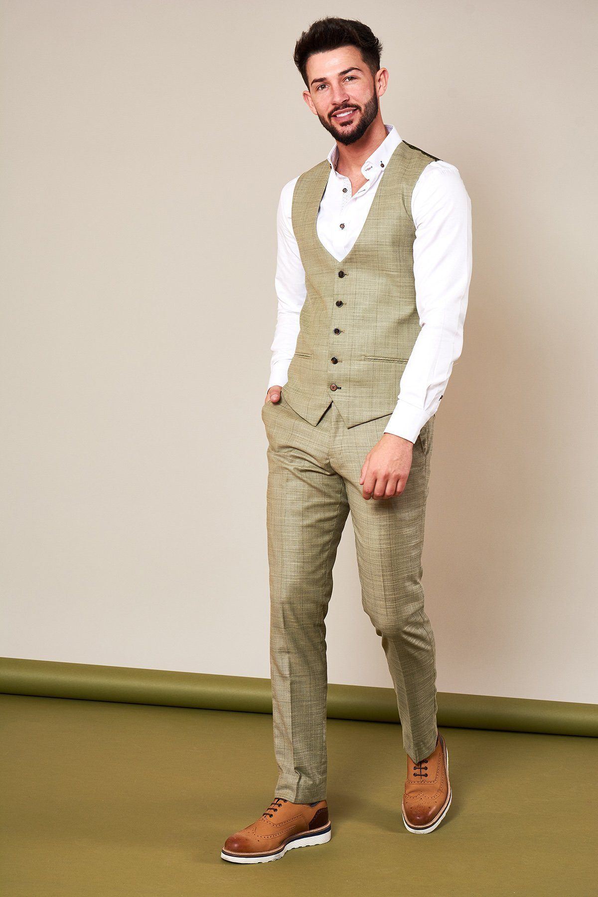A bearded man smiles in the Marc Darcy SID - Mustard Beige Check Single Breasted Waistcoat with matching slim fit pants, white shirt, and brown dress shoes, standing with one hand in his pocket against a beige background.