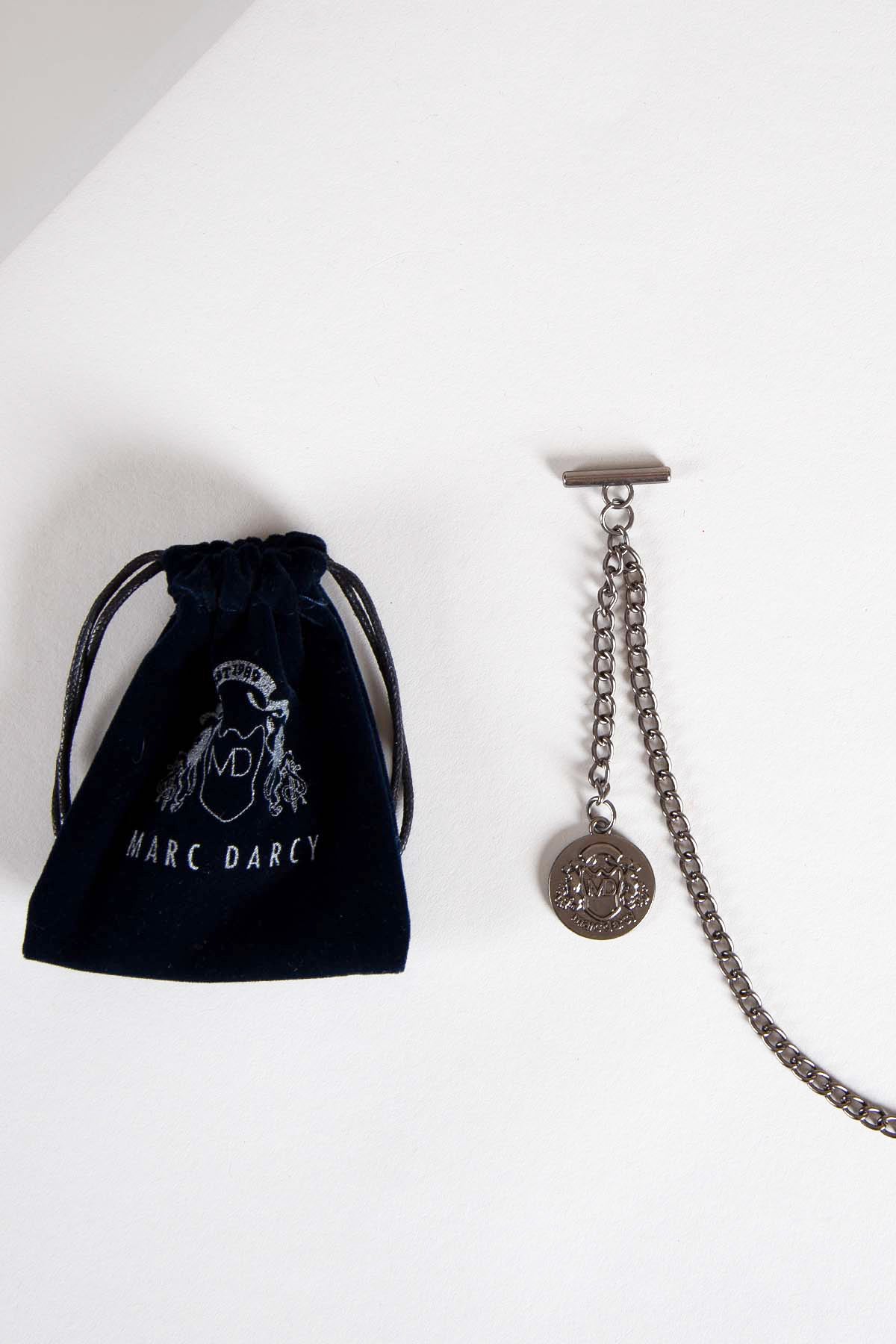 An Antique Style Silver Plated Marc Darcy T-Bar Waistcoat Chain with a coin charm comes with a dark blue velvet keepsafe pouch embroidered in silver, both displayed on a white surface.
