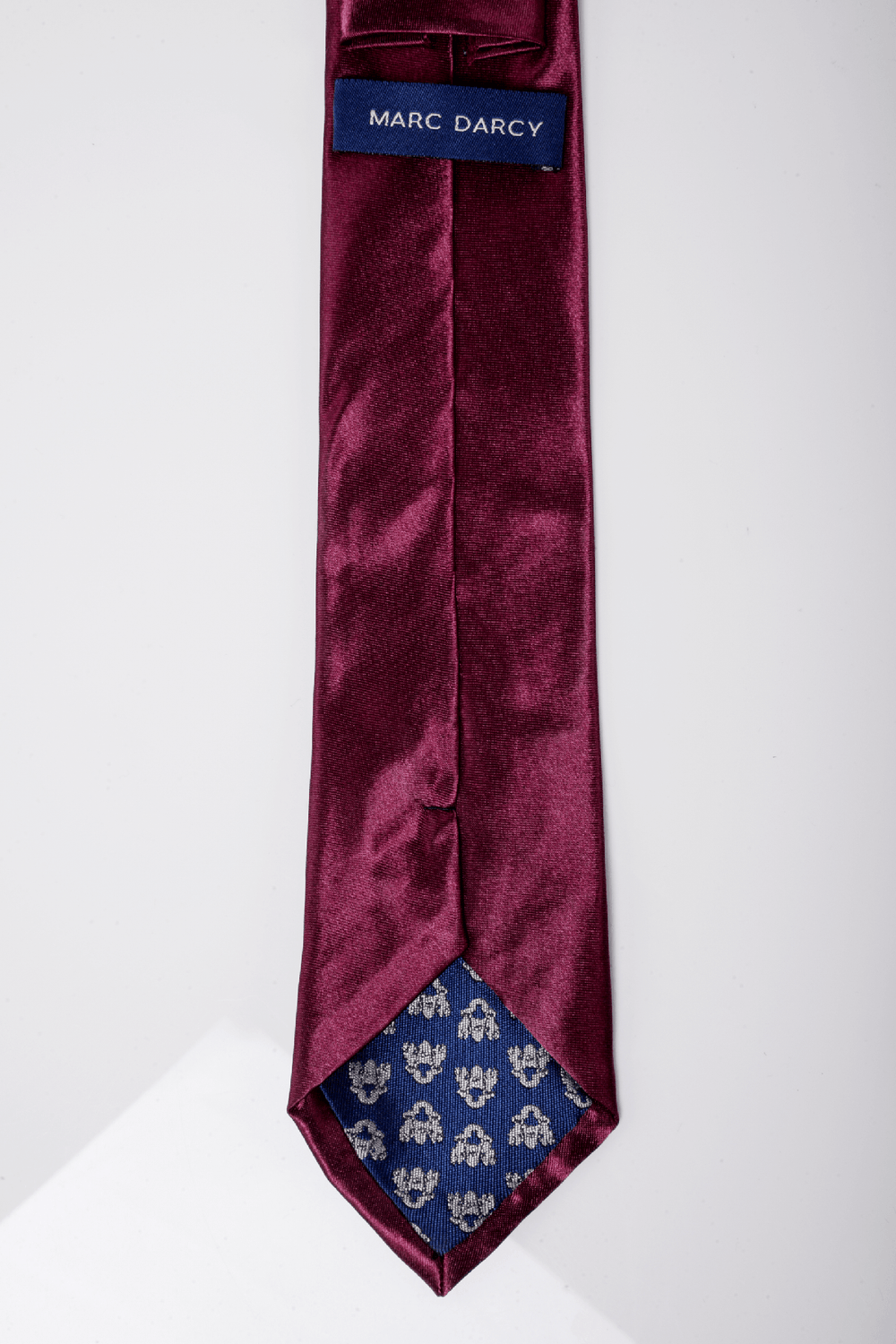 A close-up of the Accessory ST Satin Tie and Pocket Square Set in plum. The tie has a blue fabric label and tip lined with blue fabric featuring a small white floral pattern, perfect for elevating formal looks.