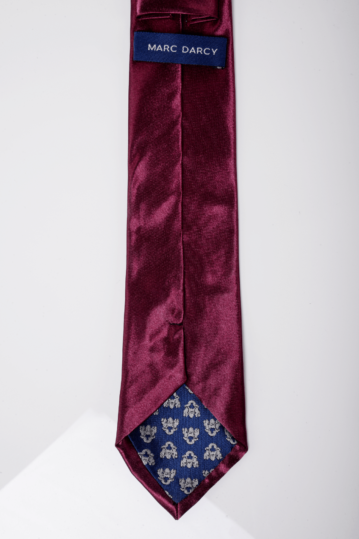 A close-up of the Accessory ST Satin Tie and Pocket Square Set in plum. The tie has a blue fabric label and tip lined with blue fabric featuring a small white floral pattern, perfect for elevating formal looks.