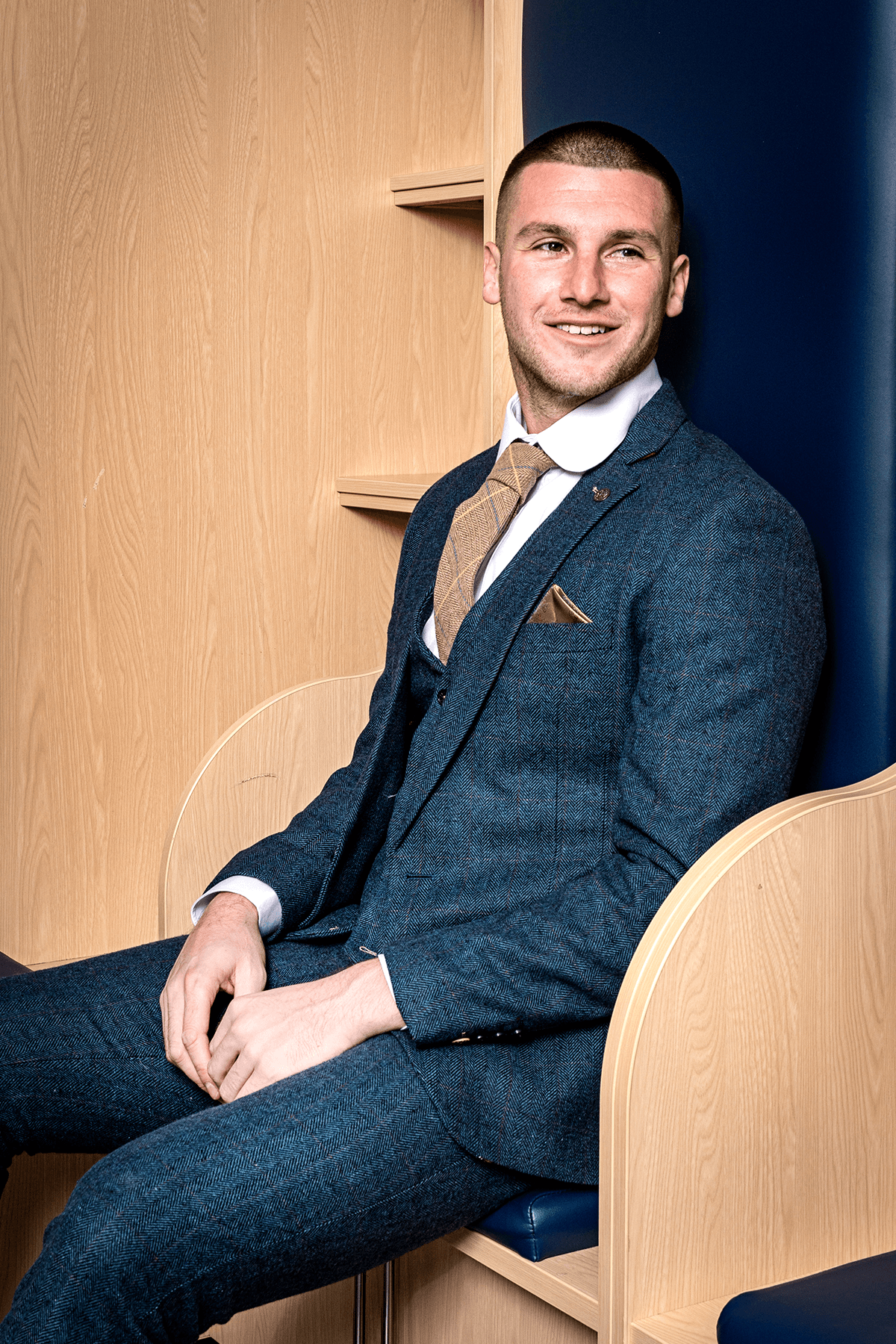A man in the marcdarcy DION Blue Tweed Suit as worn by Sam Johnstone smiles and looks to the right, seated in a wooden booth with blue accents. He pairs the suit with a white shirt and tan checkered tie.