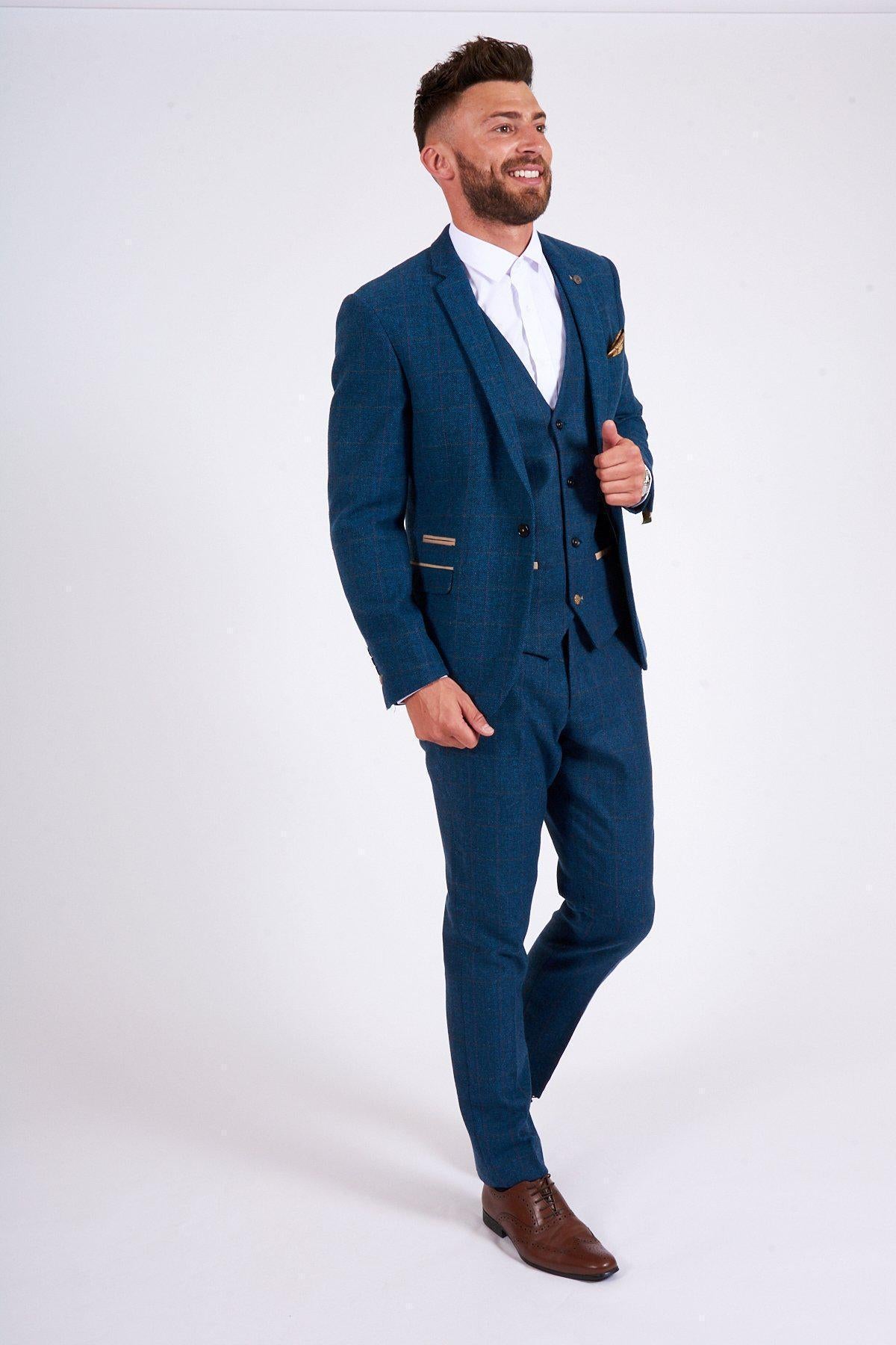 A man with a neatly trimmed beard and short hair smiles in the marcdarcy DION Blue Tweed Suit as worn by Sam Johnstone, styled with a white shirt and brown dress shoes. He wears the slim fit blazer from this heritage three piece ensemble.
