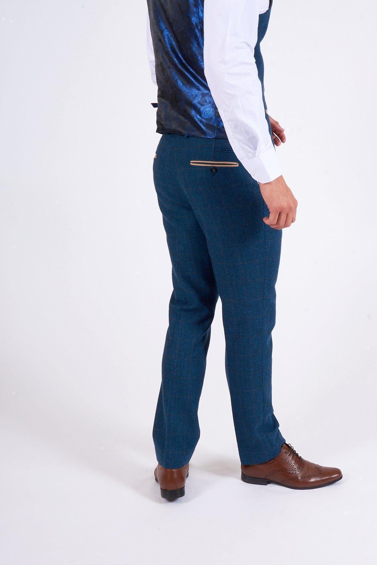 A man in a marcdarcy DION Blue Tweed Suit as worn by Sam Johnstone, brown dress shoes, and a white shirt with dark blue waistcoat stands on a white background, turned away and glancing to the side.