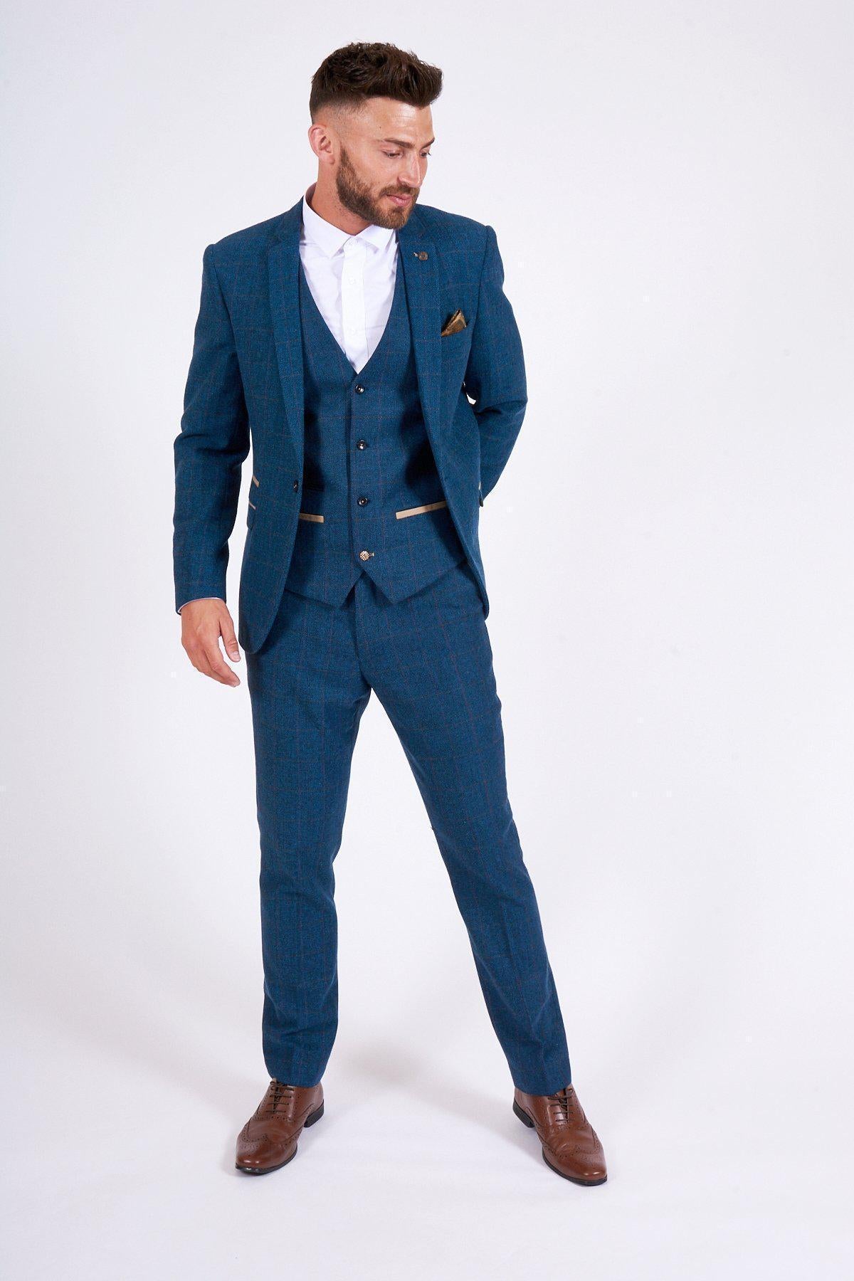 A man stands against a white background wearing the marcdarcy DION Blue Tweed Suit as worn by Sam Johnstone, paired with a white dress shirt, brown shoes, and a gold pocket square. He looks down with his hands behind his back.