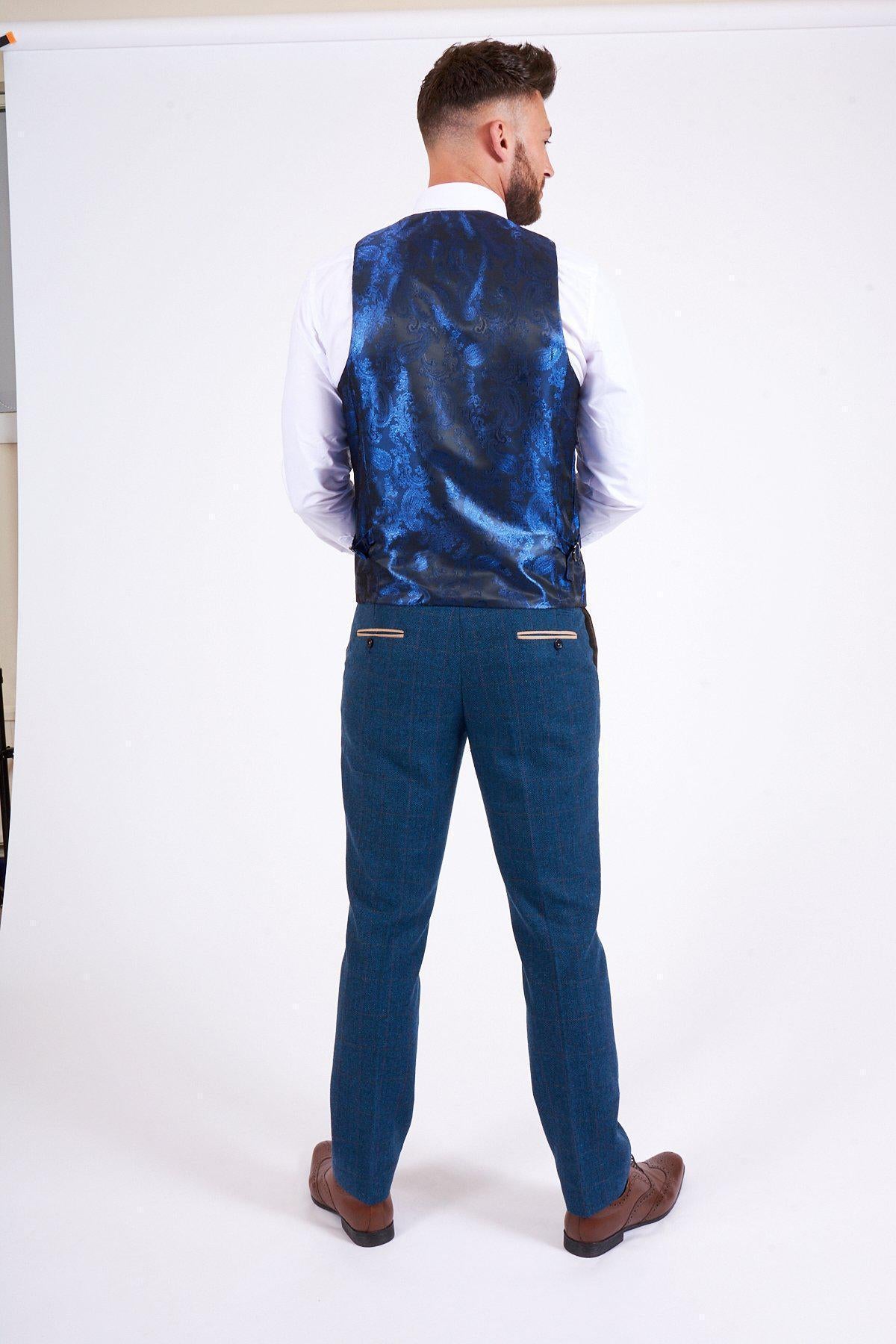 A man stands with his back to the camera wearing the marcdarcy DION Blue Tweed Suit as worn by Sam Johnstone, including a white shirt, blue patterned vest, blue trousers, and brown dress shoes against a plain white background.