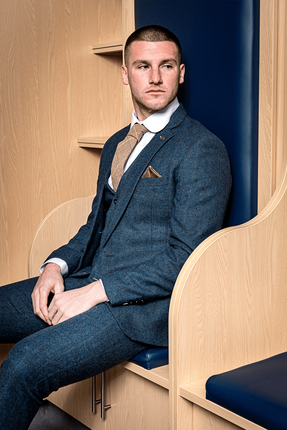 A man in the marcdarcy DION Blue Tweed Suit as worn by Sam Johnstone, paired with a brown tie and pocket square, sits on a wooden bench with blue cushions, looking left with a serious expression.