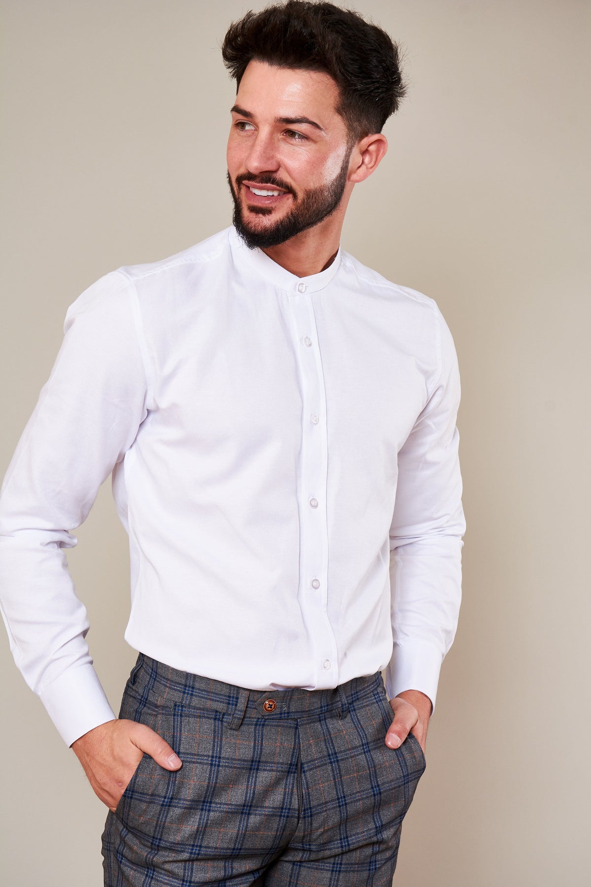 A man with dark hair and a beard smiles, wearing the Marc Darcy THOMAS White Grandad Collar Shirt and plaid pants. His hands are in his pockets, highlighting a smart-casual look.