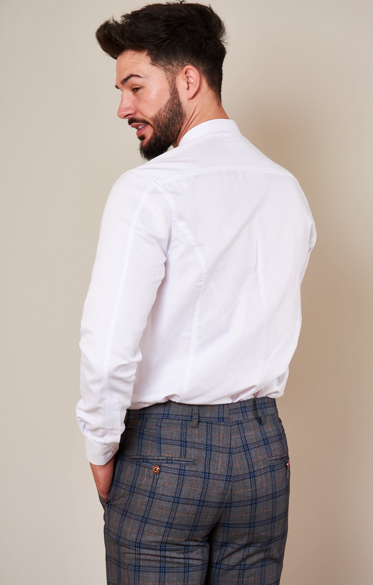 A man with short dark hair and a beard stands sideways, looking over his shoulder. He wears the Marc Darcy THOMAS White Grandad Collar Shirt and gray plaid trousers with his hands in his pockets against a plain light background.