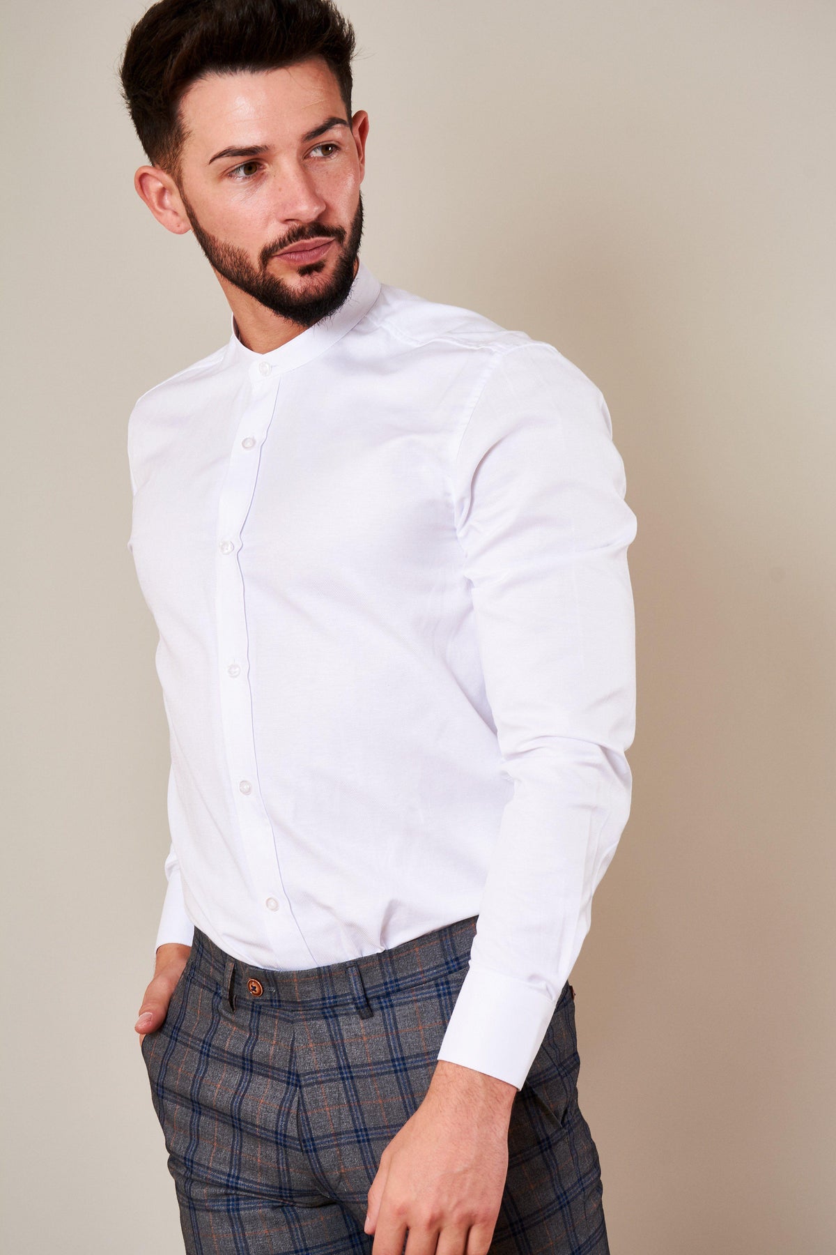 A man with short dark hair and a beard wears the Marc Darcy THOMAS White Grandad Collar Shirt and gray plaid pants, standing against a plain light background and looking to his left.