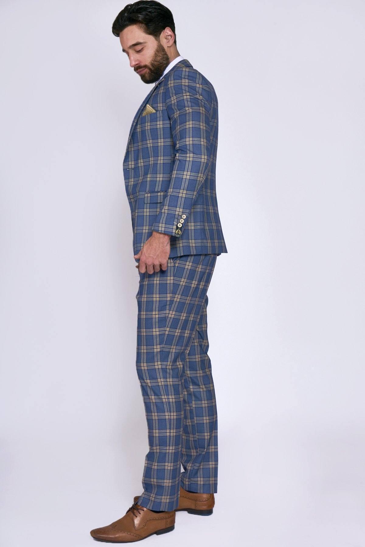 A man stands in profile wearing the marcdarcy TONY Blue Yellow Check Suit with a single breasted waistcoat, paired with a white shirt and brown dress shoes, against a plain light background.