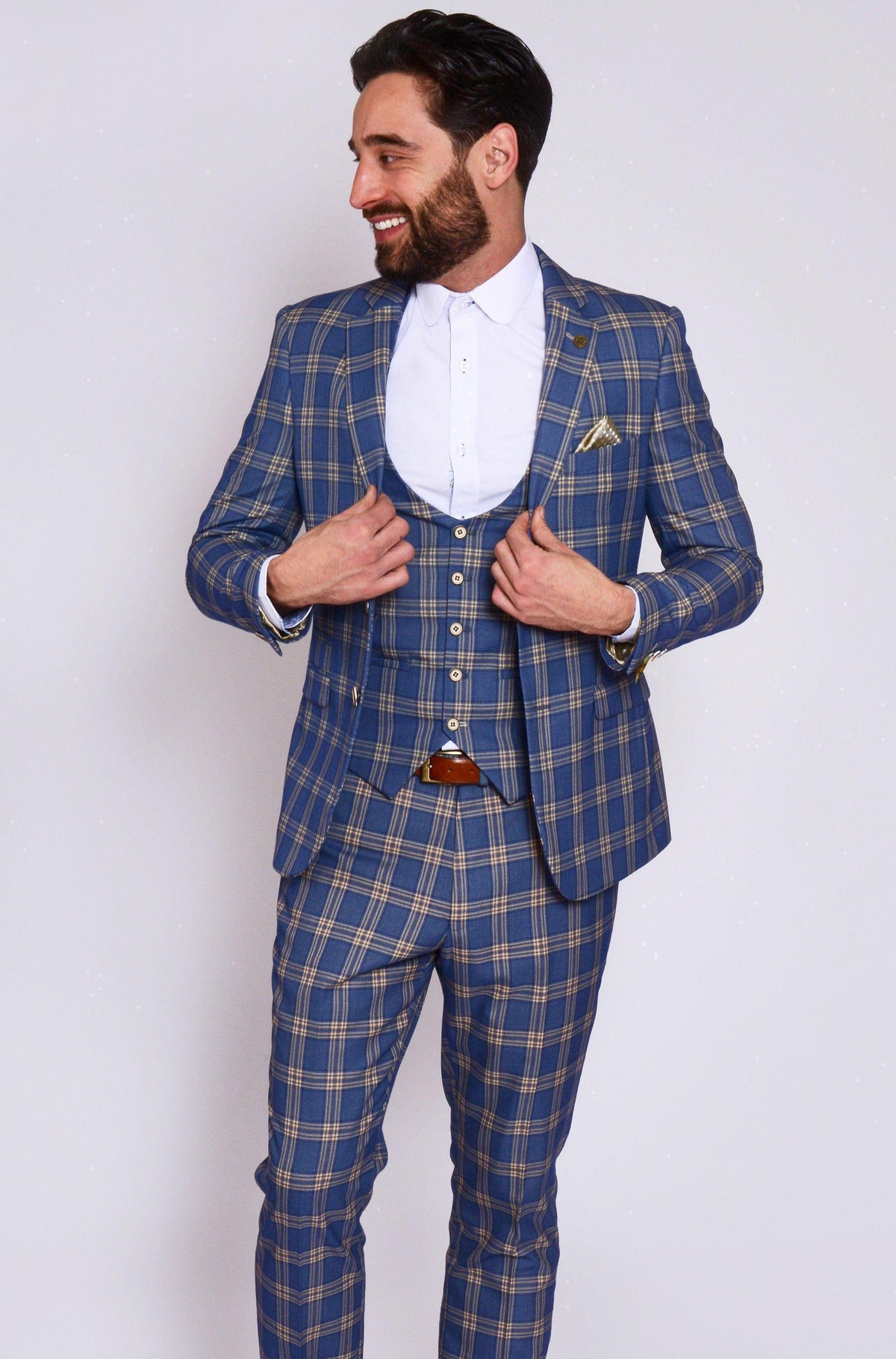 A bearded man smiles in the marcdarcy TONY - Blue Yellow Check Suit with Single Breasted Waistcoat, styled with a white shirt, brown belt, and pocket square, posing against a plain light background.