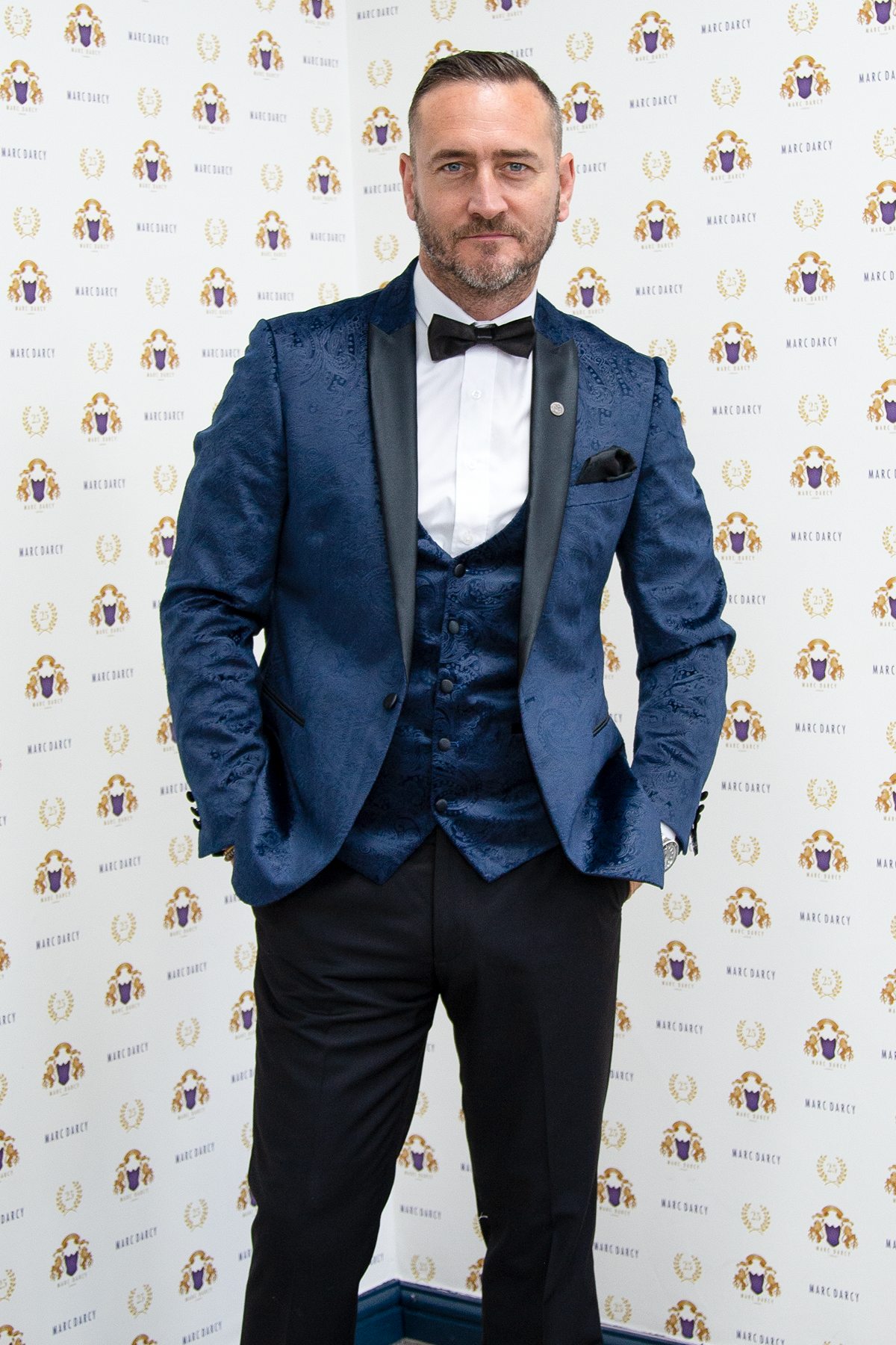 Will Mellor (Harvey Gaskell from Coronation Street) poses confidently in a Marc Darcy Simon Velvet Blazer & Waistcoat, in front of a backdrop featuring repeating Marc Darcy logos.