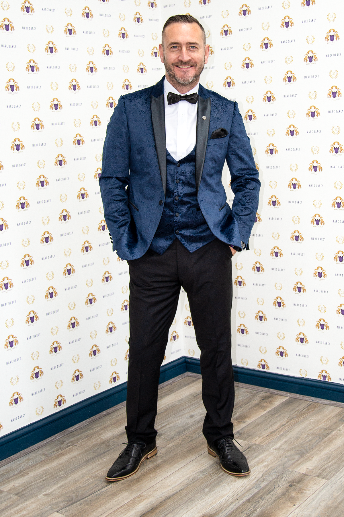 Will Mellor (Harvey Gaskell from Coronation Street) wears a Marc Darcy Simon Velvet Blazer & Waistcoat, smiling on a wood floor before a backdrop featuring a repeated logo pattern.