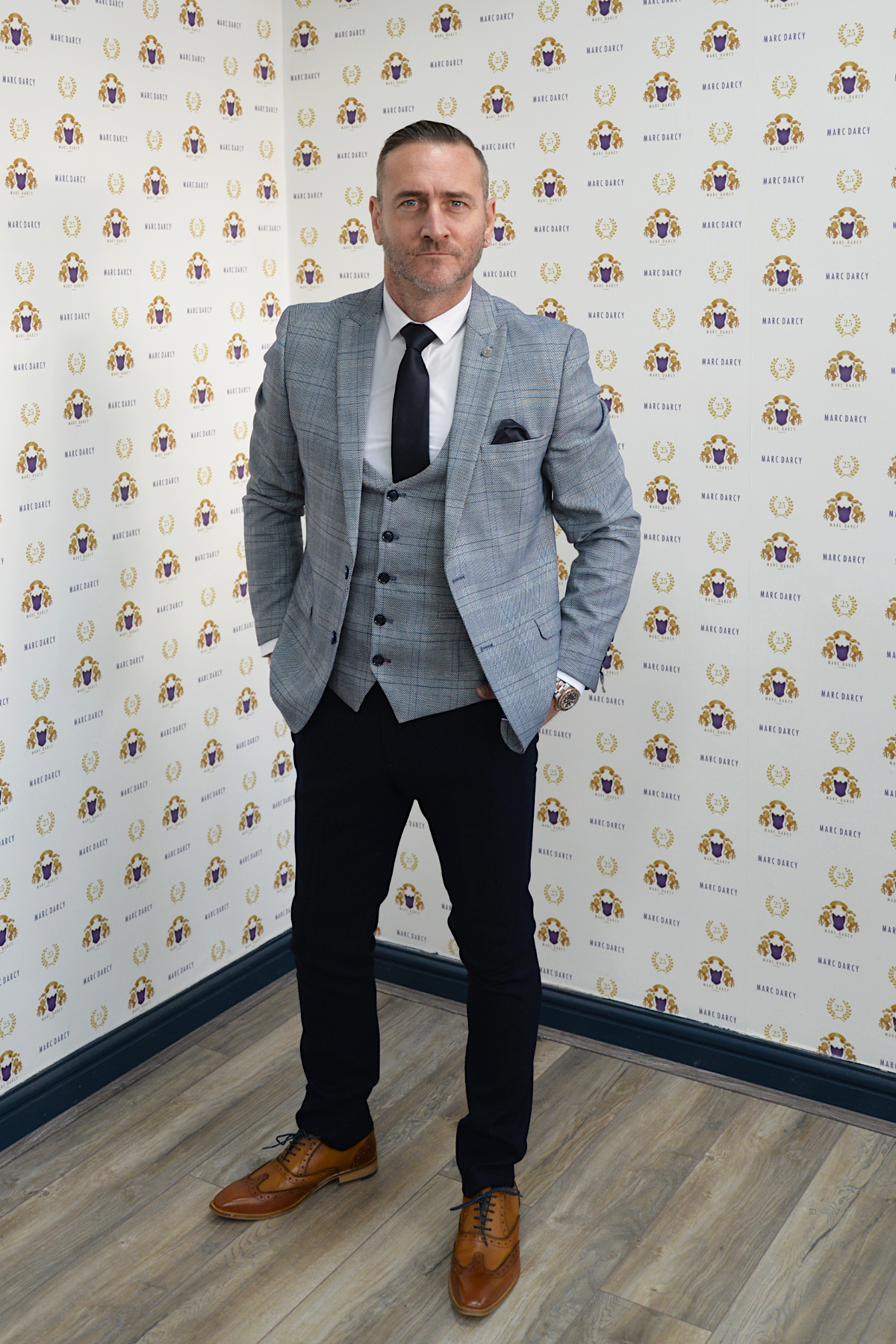 Actor Will Mellor stands on a wooden floor before a logo wall, wearing the marcdarcy Jerry Grey Check Blazer & Waistcoat. With a white shirt, black tie, and trousers, he exemplifies modern menswear style while looking confidently at the camera.