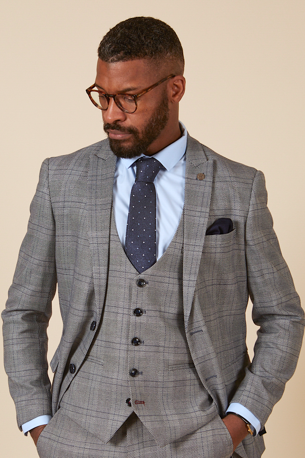 Actor Will Mellor wears a marcdarcy Jerry Grey Check Blazer & Waistcoat, styled with a light blue shirt, navy polka dot tie, and glasses. He stands hands-in-pocket, looking down to his right against a plain background.