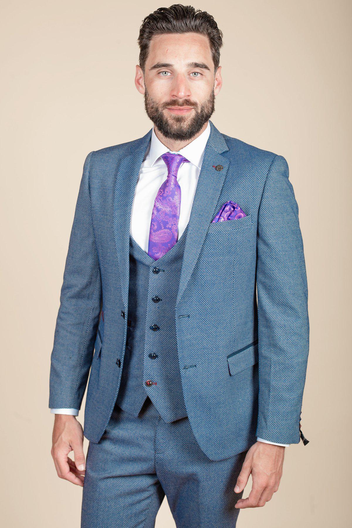 A man with dark hair and a beard wears the marcdarcy WILSON - Blue Birdseye Blazer, styled with a white shirt and purple patterned tie and pocket square, standing against a plain beige background.