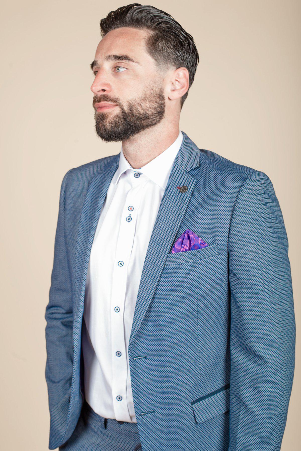 A bearded man wears the marcdarcy WILSON - Blue Birdseye Blazer in a slim fit, styled with a white shirt and purple pocket square, standing against a beige background and looking to the side.