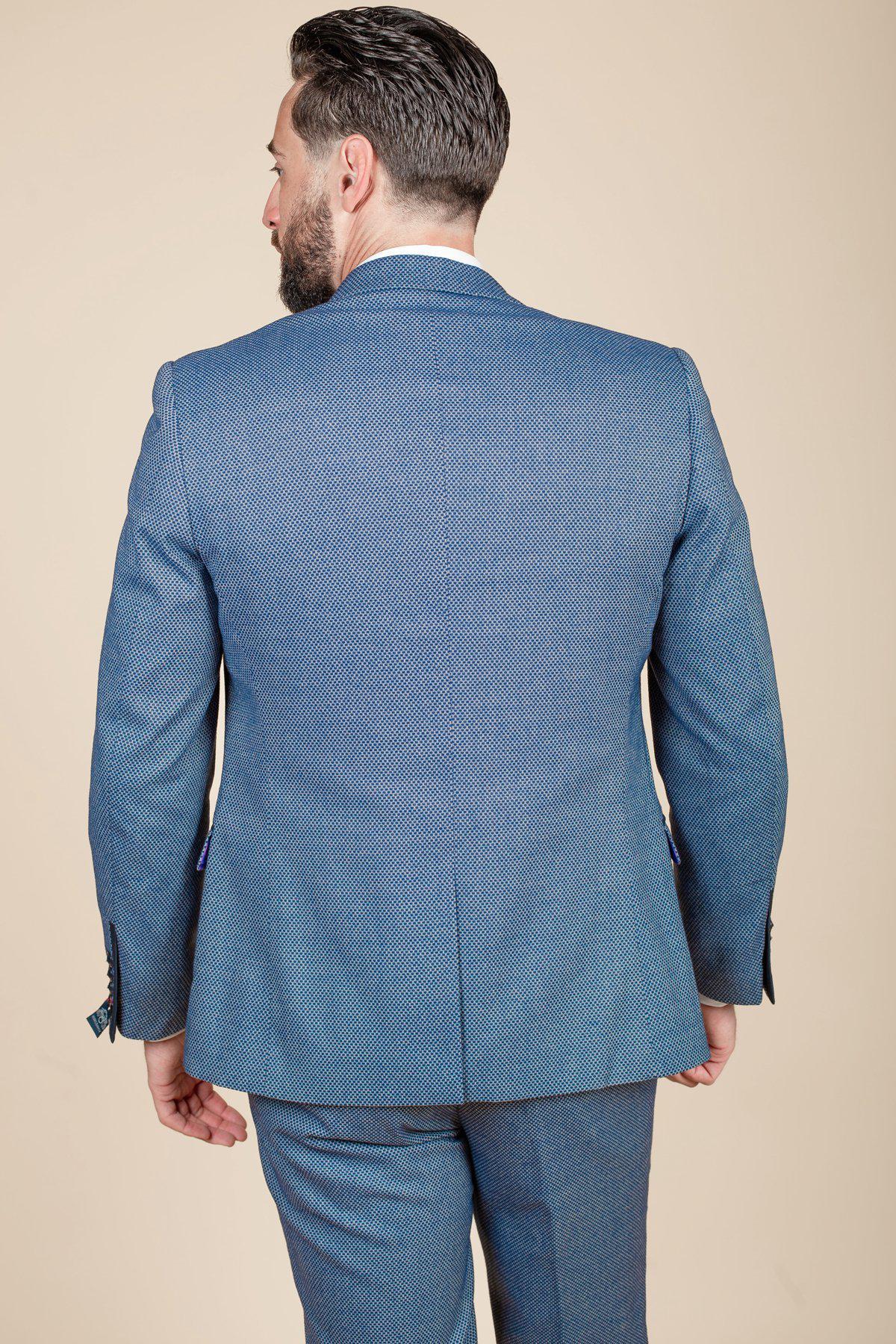 A man with short dark hair and a beard is seen from behind, wearing the marcdarcy WILSON - Blue Birdseye Blazer with matching trousers, standing against a plain beige background.
