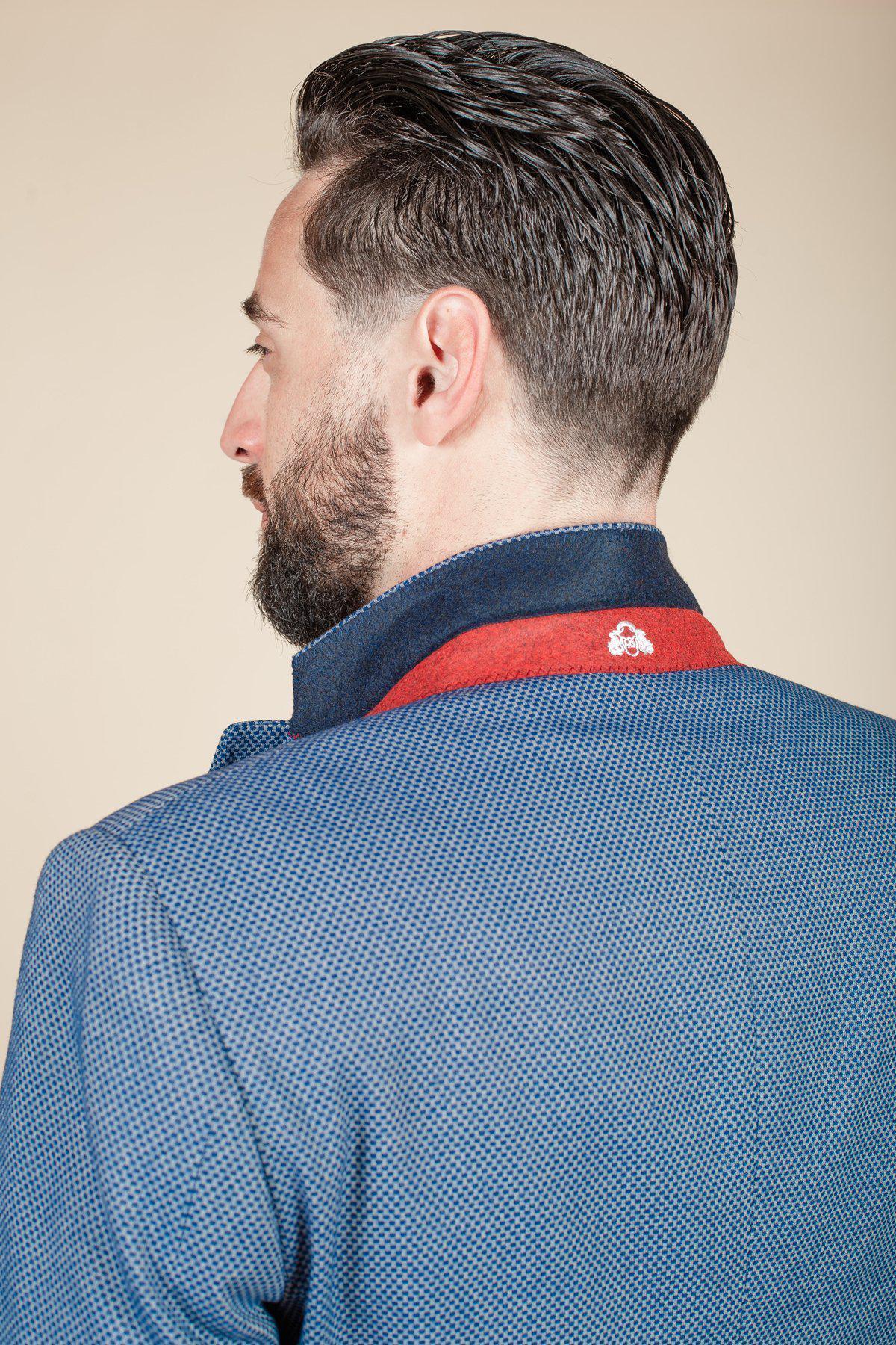 A man with dark, slicked-back hair and a beard is seen from behind wearing the marcdarcy WILSON - Blue Birdseye Blazer, featuring a slim fit, raised collar with red lining, and a small white emblem.