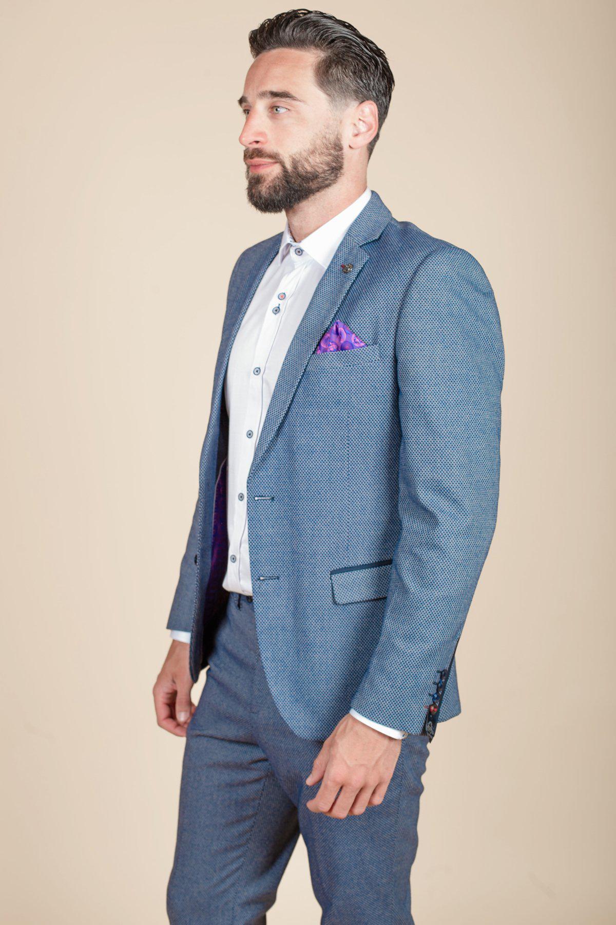 A bearded man with slicked-back hair stands sideways in a marcdarcy WILSON - Blue Birdseye Blazer, paired with a white dress shirt and purple pocket square, set against a plain beige background.