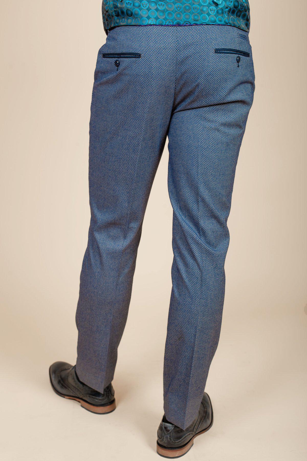 A person faces away from the camera, wearing marcdarcys WILSON Blue Birdseye Trousers, a teal patterned shirt, and black dress shoes against a light background.