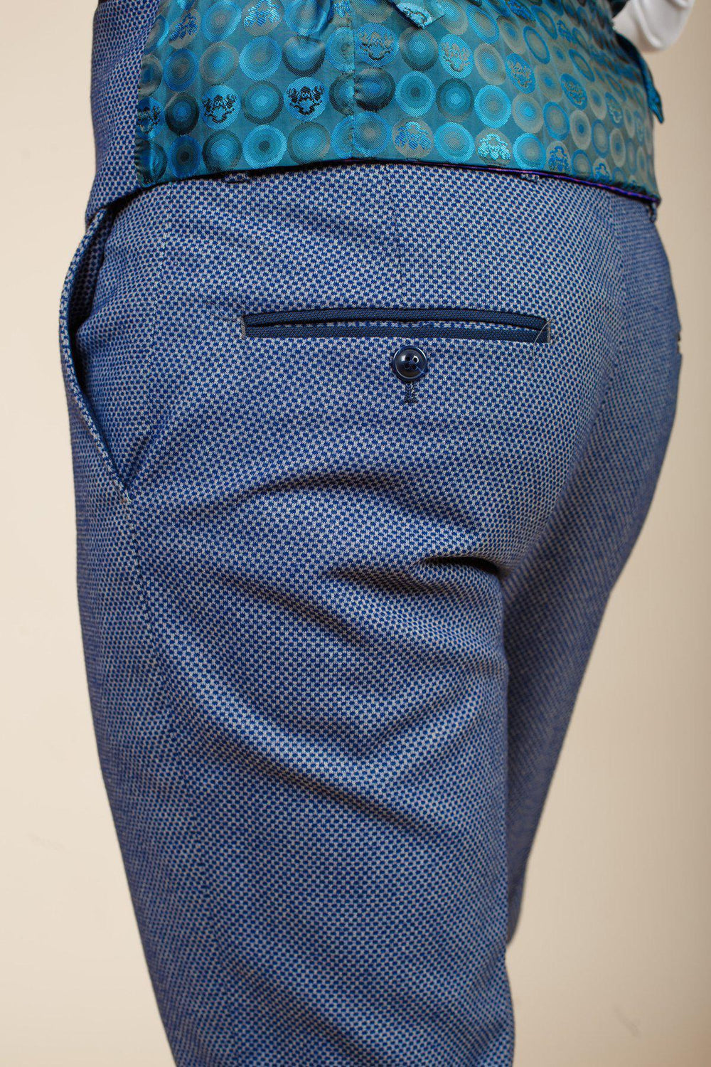 Close-up of a person wearing marcdarcy WILSON - Blue Birdseye Trousers with a slim fit, buttoned back pocket, and a waistband featuring a blue and teal circular pattern. Set against a plain beige background.