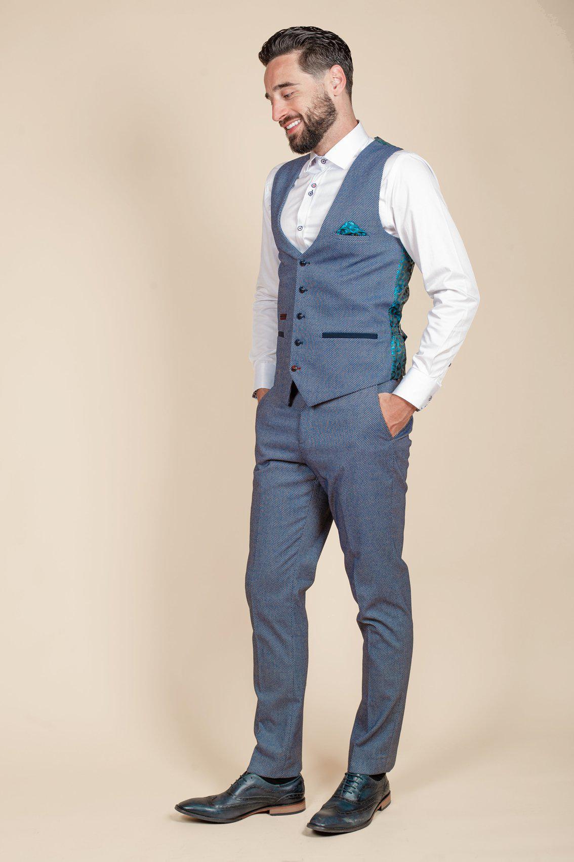A man with dark hair and a beard smiles in profile against a beige background, wearing marcdarcy WILSON Blue Birdseye Trousers, a matching light blue three-piece suit, a white shirt, and black dress shoes—no tie.