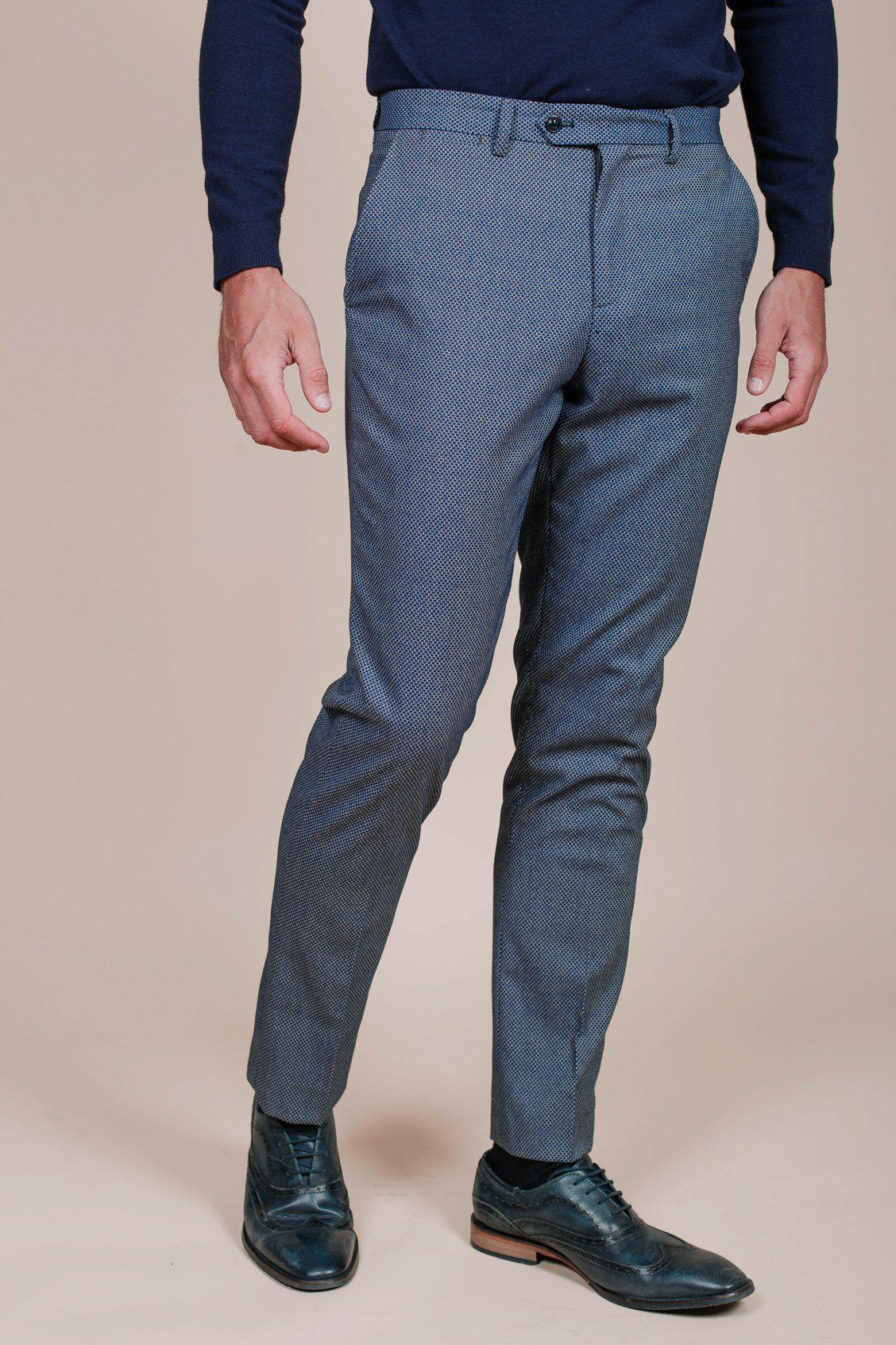 A person wears marcdarcy WILSON - Blue Birdseye Trousers with a navy long-sleeve shirt and dark blue leather dress shoes, standing against a plain beige background.