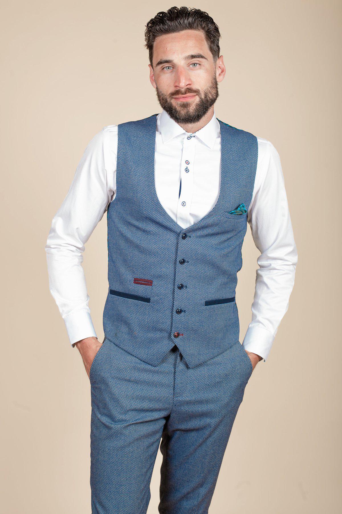 A man with dark hair and a beard wears the marcdarcy WILSON Blue Birdseye Waistcoat and matching trousers over a white shirt, standing with hands in pockets against a beige background and looking at the camera with a slight smile.