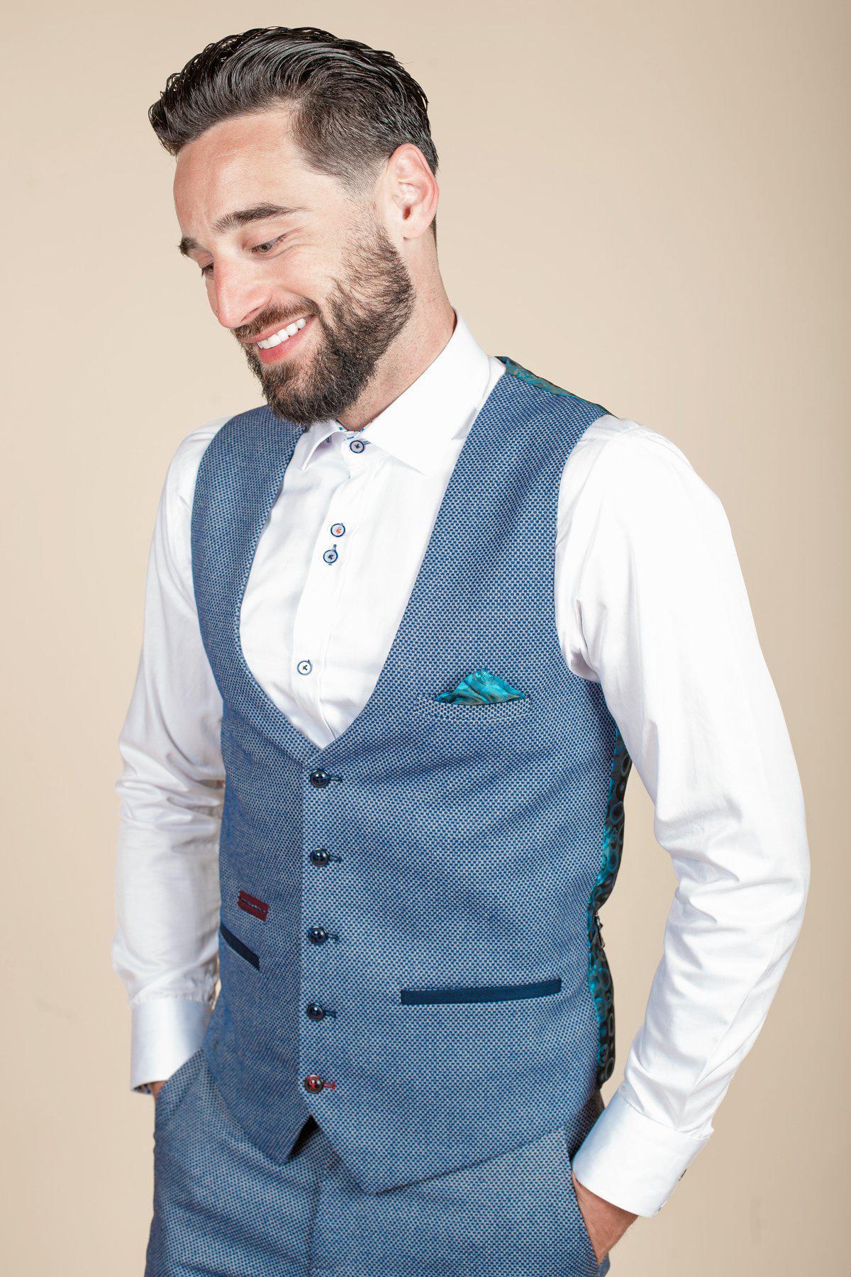 A bearded man with dark hair smiles, looking down with his hands in his pockets. He wears the marcdarcy WILSON - Blue Birdseye Waistcoat over a white shirt, styled with matching pants and a teal pocket square, set against a beige background.