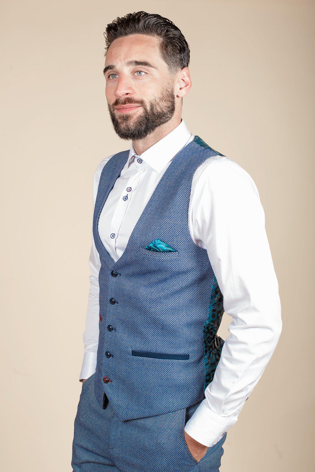 A man with short dark hair and a beard wears the marcdarcy WILSON - Blue Birdseye Waistcoat with matching trousers, a white dress shirt, and a teal pocket square, looking to the side against a plain beige background.